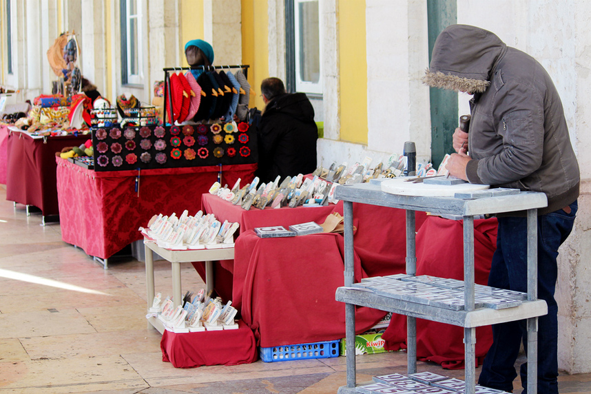 Handmade goods for sale and a merchant carver crafting wares at the Praca do Comercio handicraft market, Lisbon, Portugal.