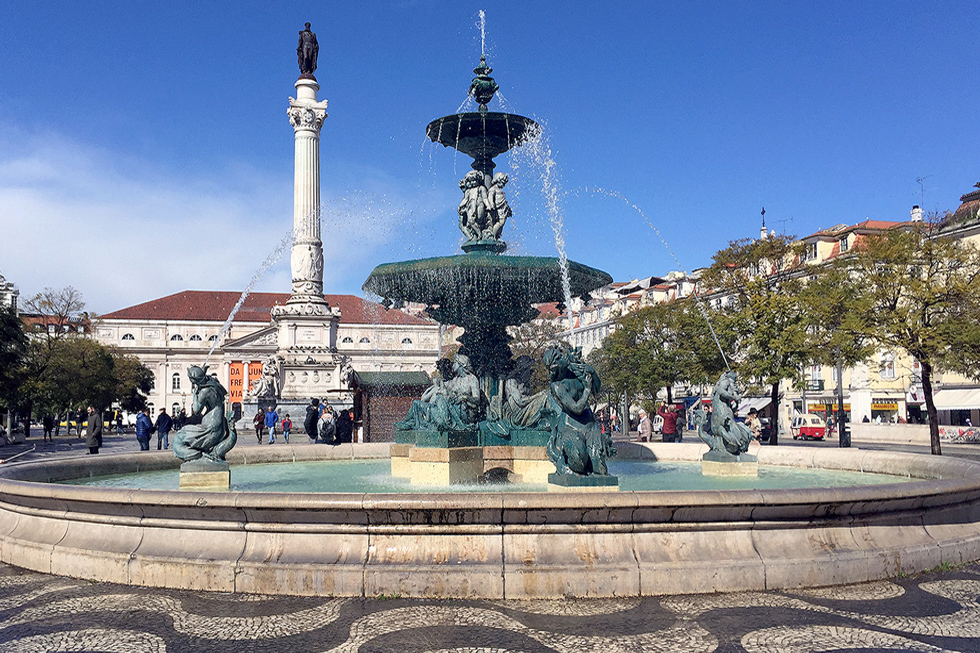 A bronze fountain on the south side of the square with the column of Pedro IV and The National Theatre D.Maria II in the background. Pedro IV/ Rossio Square, Lisbon, Portugal.